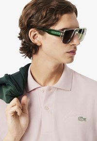 Solglasögon med överdimensionerade rektangulära linser i grönt och transparenta bågar. Bär en ljusrosa polo med broderad logotyp och en grön tröja draperad.