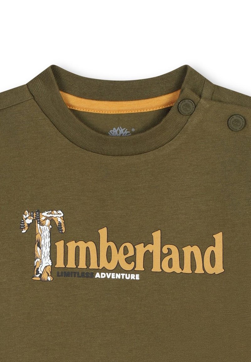 T-shirt verde oliva con scollatura tonda, due bottoni sulle spalle e logo "Timberland" in caratteri stilizzati, con un accento giallo.