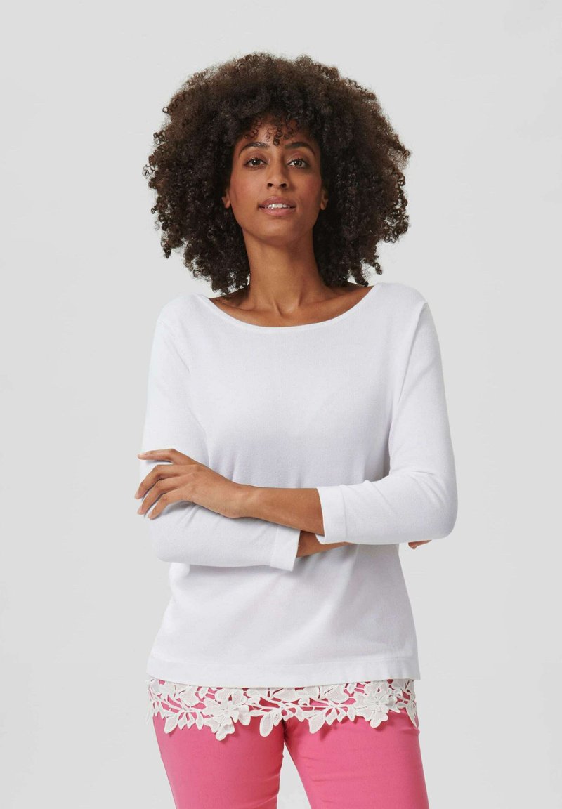 Witte lange mouwen top met een brede halslijn, versierd met een decoratieve witte kanten rand aan de onderkant. Gewaard met lichtroze broek.