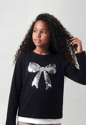 Fille aux longs cheveux bouclés portant un t-shirt noir à manches longues avec un grand nœud en sequins argentés, touchant ses cheveux et regardant sur le côté.
