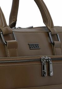 Braune Lederhandtasche mit doppelten Reißverschlüssen, silberner Hardware und einem schwarzen Logo-Patch. Das Design umfasst strukturierte Griffe und Stichdetails.