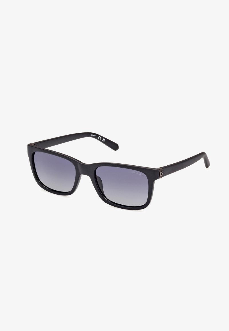 Lunettes de soleil carrées noires avec des verres dégradés, présentant une finition mate et un logo subtil sur la branche. Design de monture en plastique léger.