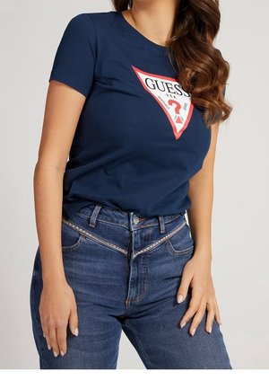 Tummansininen puuvillainen t-paita, jossa edessä punainen kolmionmuotoinen "GUESS"-logo. Yhdistetty korkeavyötäröisiin sinisiin farkkuihin.