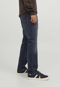 Jeans in denim blu scuro con taglio a gamba dritta, dotati di tasche posteriori e sbiaditure leggere, abbinati a sneakers blu navy con dettagli bianchi.