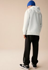 Haut de survêtement gris clair avec capuche, associé à un pantalon de survêtement noir et des baskets noires. Bonnet en laine bleu ; présente un texte blanc sur la jambe du pantalon.
