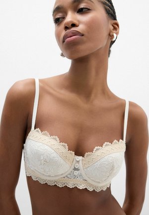 Mujer con sujetador de encaje blanco con tirantes finos y detalle de botones, mirando ligeramente de lado sobre un fondo blanco liso.