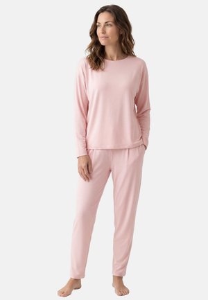 Vrouw staat blootsvoets, draagt een lichtroze top met lange mouwen en broek, kijkt iets opzij tegen een effen witte achtergrond.