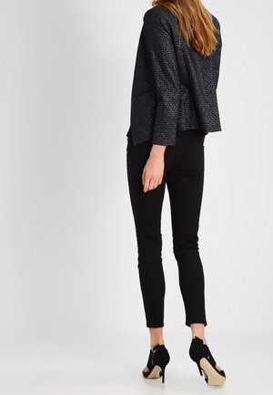 Mujer de espaldas, vestida con una prenda superior negra texturizada de manga larga, pantalones negros ajustados y zapatos negros de tacón alto.