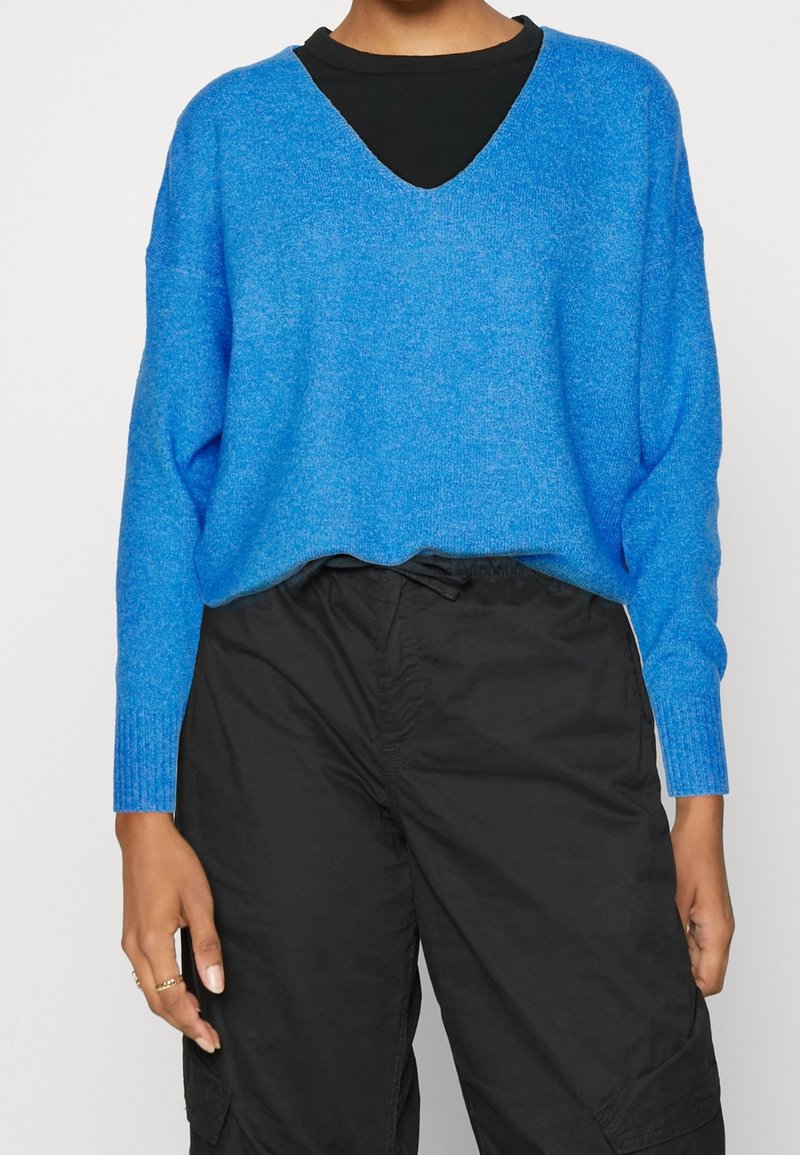 Personne portant un pull bleu vif à col en V par-dessus une chemise noire avec un pantalon noir, les mains détendues le long du corps.