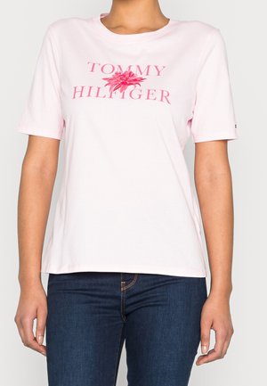 Kobieta nosząca jasnoróżowy T-shirt z krótkim rękawem z napisem „Tommy Hilfiger” i kwiatowym motywem, zestawiony z ciemnoniebieskimi dżinsami.