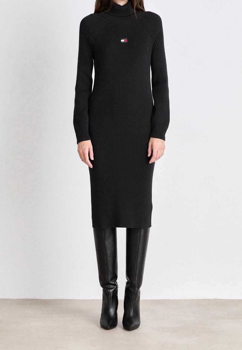 Femme portant une robe midi en maille côtelée noire à manches longues avec un petit logo rouge, blanc et bleu sur la poitrine, associée à des bottes noires montantes.