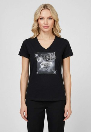 Donna con capelli biondi indossa una maglietta nera con scollo a V e grafica argentata scintillante con la scritta "United Artists" in un design quadrato, abbinata a pantaloni neri.