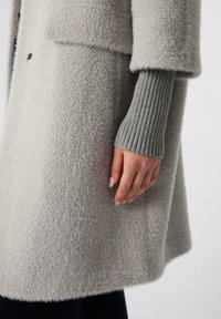 Persona che indossa un cappotto in lana grigia chiara con una texture, con lunghi polsini a coste grigi che mostrano la mano e parte della manica e della parte anteriore del cappotto.