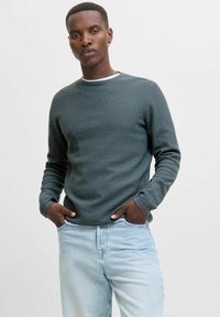 Homme aux cheveux courts portant un pull texturé vert foncé et un jean bleu clair, se tenant avec les mains dans les poches sur un fond clair et uni.