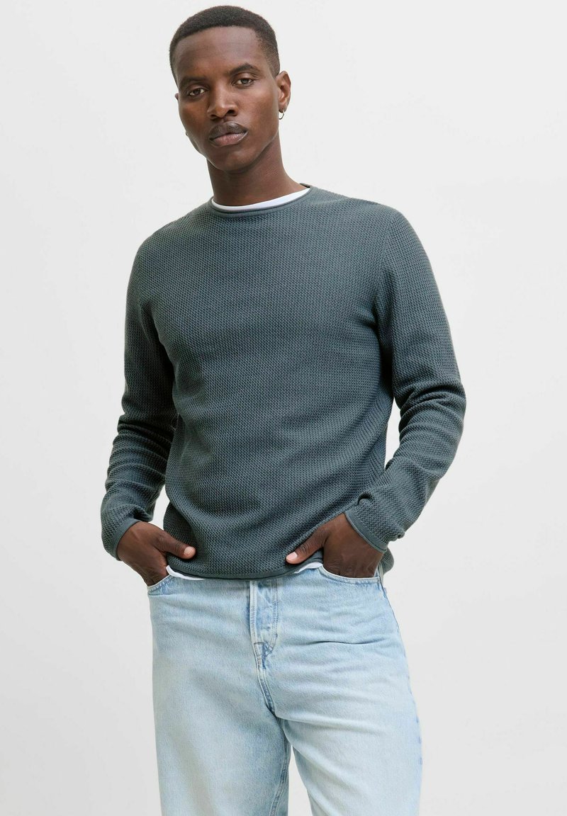 Homme aux cheveux courts portant un pull texturé vert foncé et un jean bleu clair, se tenant avec les mains dans les poches sur un fond clair et uni.