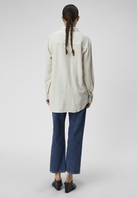 Chemise beige clair à manches longues avec un col, présentant un ourlet arrondi. Associée à un jean en denim bleu à jambes larges et à des mules noires à talons.