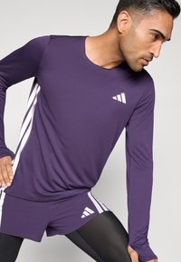 Lila langärmliges Sportshirt mit weißen Seitenstreifen und Logo. Shorts und schwarze Leggings vervollständigen das sportliche Outfit. Glatte Textur.