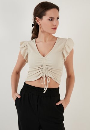 Kremasti rebrasti crop top s dubokim V-izrezom, nabranim prednjim dijelom, vezanjem na užad i kratkim volanastim rukavima, u kombinaciji s crnim hlačama s elastičnim pojasom.