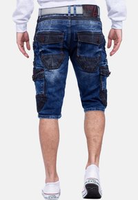 Homme portant un short en denim bleu cargo avec plusieurs poches et des baskets Converse All Star blanches, vu de dos.