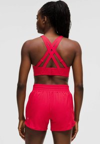 Sujetador deportivo rojo con tirantes cruzados y broche, combinado con pantalones cortos rojos de cintura elástica. Tejido suave y ligero, con un detalle mínimo de logotipo.