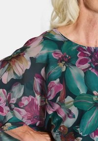 Bloes met bloemenprint met grote roze en groene bloemen op een donkere achtergrond, gemaakt van lichtgewicht, doorschijnende stof met een ronde hals.
