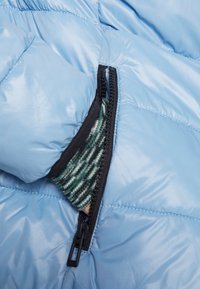 Doudoune bleu clair avec une fermeture éclair noire, révélant une doublure rayée verte et blanche dans la poche partiellement ouverte. Texture lisse.