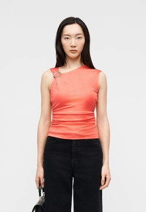 Femme aux longs cheveux foncés portant un haut corail sans manches avec une boucle argentée sur une épaule et un pantalon noir, tenant un sac à main noir.