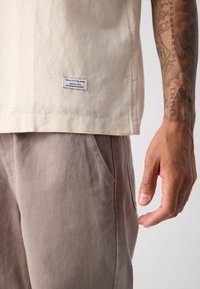 Beige bavlněná košile s krátkými rukávy a štítkem s nápisem "Marco Polo Denim." Šedohnědé kalhoty mají boční kapsu a texturovanou látku.