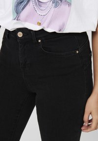 Gros plan sur un jean en denim noir avec poches avant et bouton métallique, associé à un t-shirt blanc présentant un motif d'une personne portant des colliers superposés.