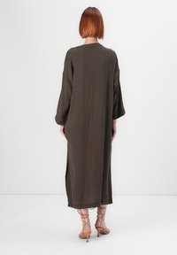 Femme aux cheveux roux courts portant une longue robe brune en tissu texturé et des escarpins beiges à lacets, se tenant contre un fond blanc uni.