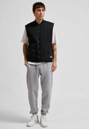 Sort polstret vest med lynlås foran og to forlommer, båret over en hvid t-shirt og grå sweatpants. Modellen er stylet med hvide sneakers.