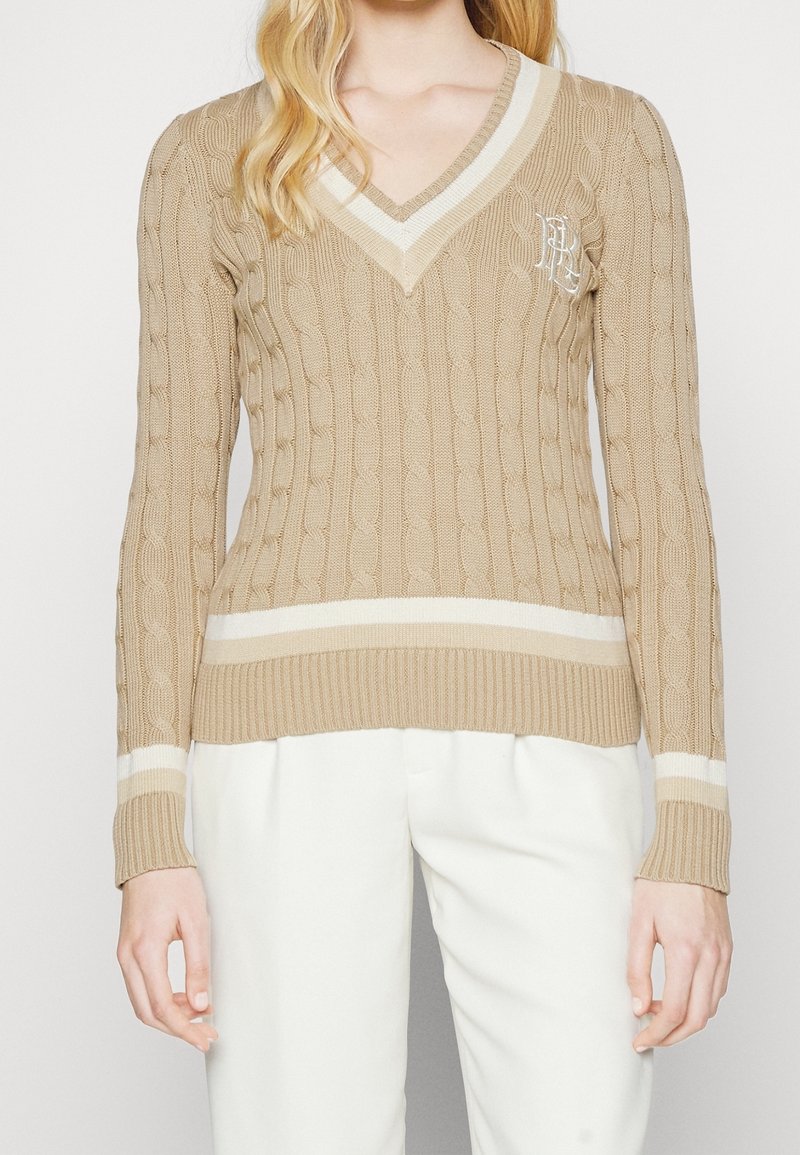 Beige stickad tröja med V-ringning, flätmönster och ribbad nederkant och manschetter med vita randdetaljer. Broderad logotyp på bröstet.