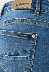 Jeans en denim bleu avec une étiquette en cuir portant le mot "GARCIA" au-dessus de la poche. Présente des coutures contrastantes et une petite étiquette de marque sur la poche arrière.
