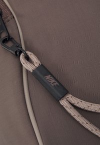 Brun tygväska med detaljer såsom ett flätat rep-handtag, en svart plastklämma och en logotypaccent på en rektangulär tag.