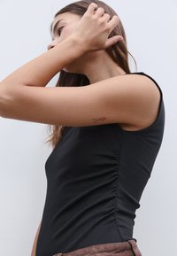 Schwarzes ärmelloses Top aus glattem Stoff mit einer gerafften Naht an der Seite. Person mit langen Haaren, die sich die Haare frisiert.