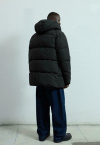 Svart puffjacka med huva, med horisontellt quiltade paneler. Bärs över löst sittande mörkblå jeans och svarta skor.