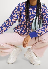 Pull bleu avec un imprimé léopard rose et blanc, associé à un pantalon large rose clair et des baskets blanches épaisses. Des accents incluent un col bleu.