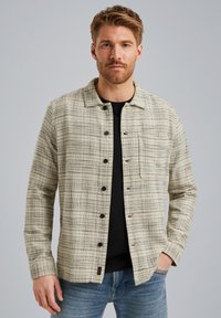 Veste en tissu à carreaux beige clair avec un col, boutons noirs, poches poitrine et manches longues, superposée sur une chemise noire.
