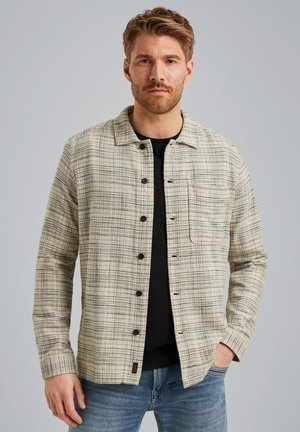Veste en tissu à carreaux beige clair avec un col, boutons noirs, poches poitrine et manches longues, superposée sur une chemise noire.