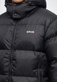 Черно подплатено яке с преден цип, висок яка с капси и бял лого Schott NYC на гърдите.