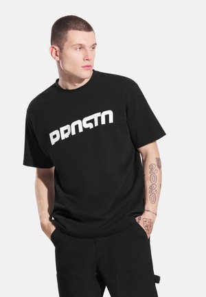 Czarny t-shirt z dużym, wyraźnym białym napisem "PRNSTN." Materiał wydaje się miękki i gładki, z krótkimi rękawami i luźnym krojem.