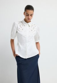 Witte blouse met opgeblazen mouwen en een knoopsluiting aan de voorkant, met ingewikkelde metalen versieringen op het schoudergedeelte. Gecombineerd met een marineblauwe rok.