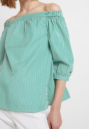 Kvinde iført en grøn og hvid vertikalt striped off-shoulder bluse med trekvartede ærmer og hvide bukser.