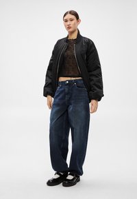 Čierna bomber bunda, skrátený hnedý top s textúrovaným vzorom, voľné modré rifle a čierne topánky s bielymi akcentmi.