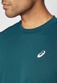 Teal kortermet treningsskjorte laget av glatt stoff, med rund hals og en hvit logo på venstre brystområde.