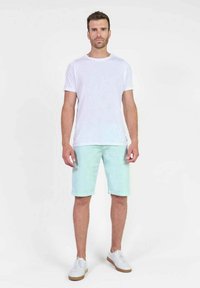Witte katoenen t-shirt, licht mintgroene shorts en witte sneakers. Model staat rechtop met de handen aan de zij, neutrale achtergrond.
