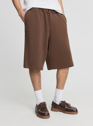 Uomo che indossa pantaloncini marroni fino al ginocchio, calzini bianchi fino a metà polpaccio e scarpe stringate in pelle marrone su uno sfondo semplice.