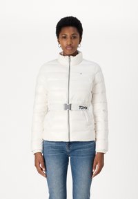 Hvit pufferjakke med høy krage og frontlås, med belte i midjen og logo-detalj. Båret over blå jeans.