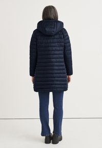 Granatowa kurtka puchowa z kapturem, z poziomymi pikowanymi sekcjami, długimi rękawami i średniej długości krojem. Noszona na ciemnych dżinsach i czarnych butach.
