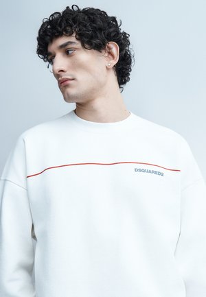Jeune homme aux cheveux bouclés et foncés portant un sweat-shirt blanc DSQUARED2 avec une ligne rouge, regardant vers sa gauche sur un fond gris clair.